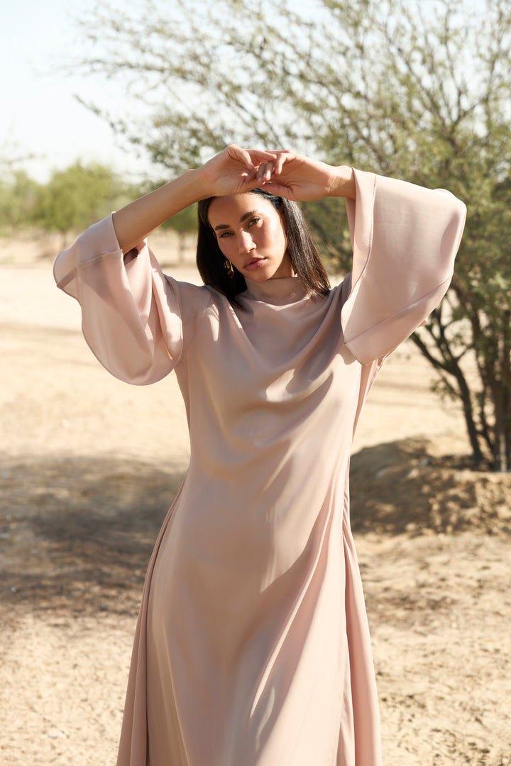 Woman in a long, light-colored dress standing in a desert-like setting with trees in the background.