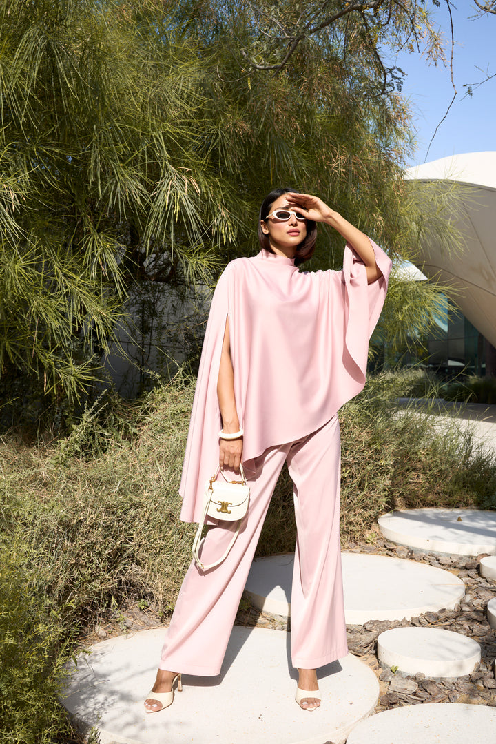 Woman in a pink outfit standing outdoors with greenery and a building in the background