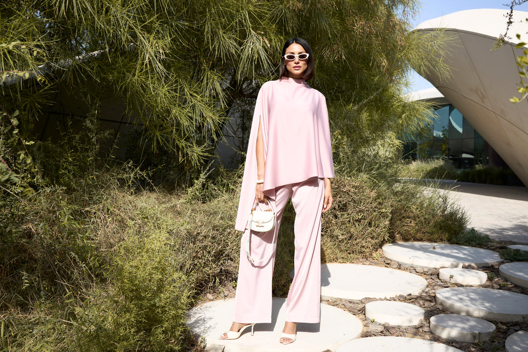 Woman in a pink outfit standing outdoors near greenery and a modern building.