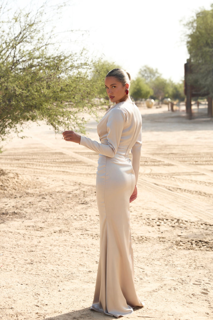 Woman in a long, light-colored dress standing in a desert-like setting with trees in the background.