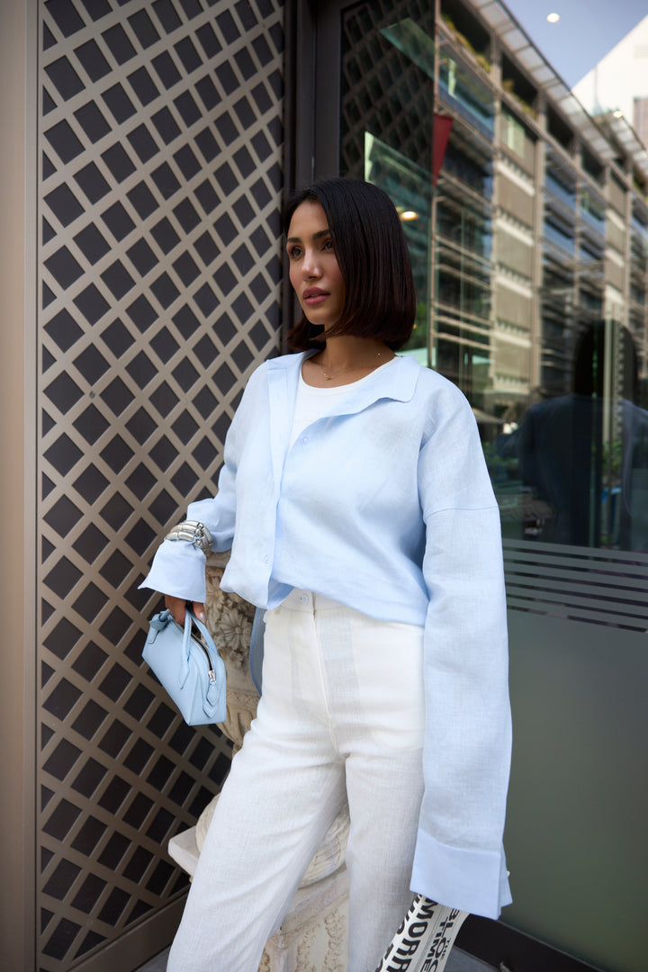 Woman in light blue jacket and white pants standing against a building with reflective glass windows.