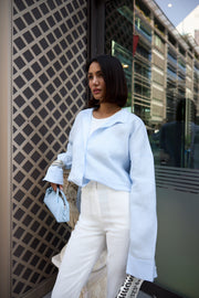 Woman in light blue jacket and white pants standing against a building with reflective glass windows.