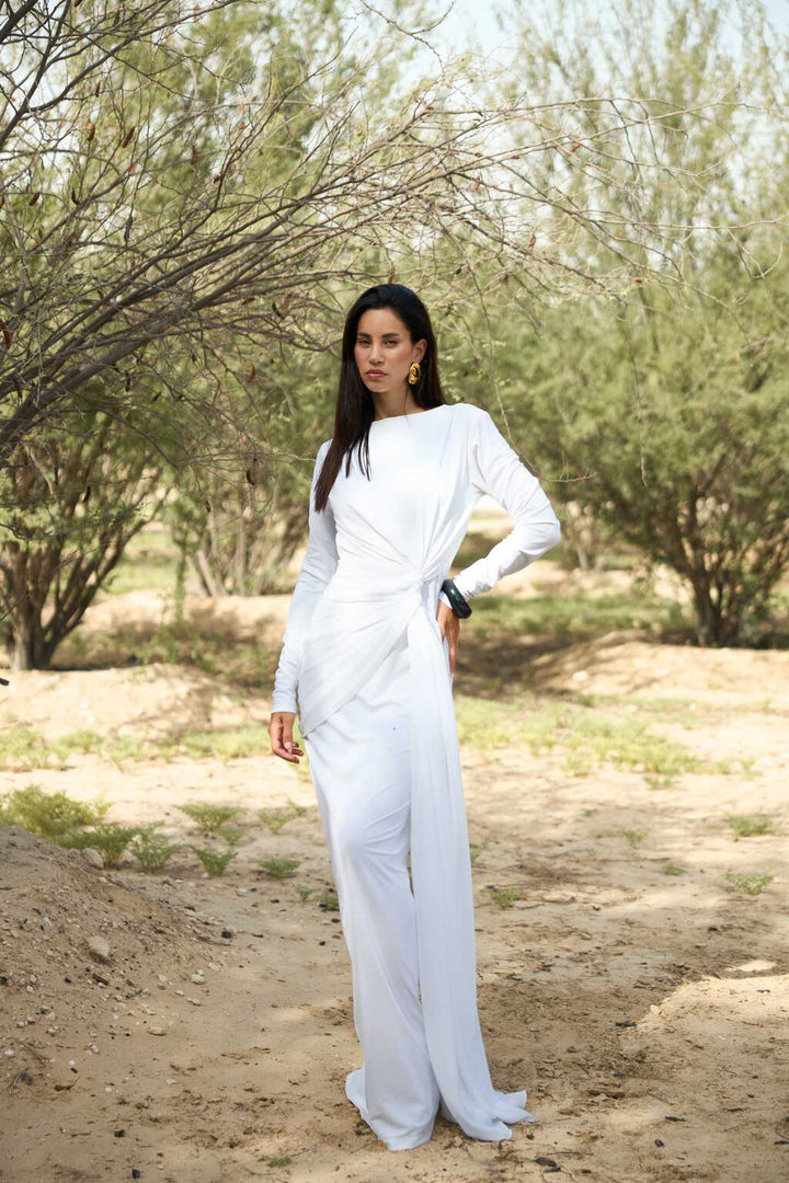 Woman in a white outfit standing in a natural setting with trees and dirt ground.