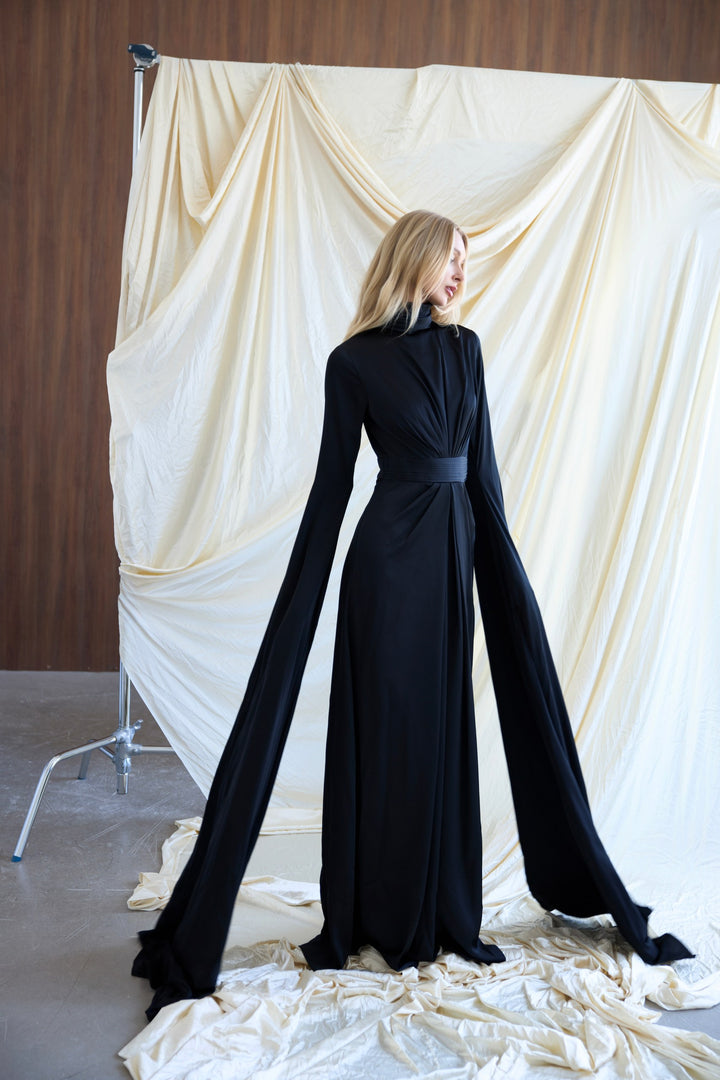 Woman wearing a black evening gown standing against a white draped curtain.