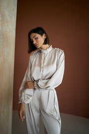 Woman wearing a light-colored blouse and pants against a warm-toned wall.